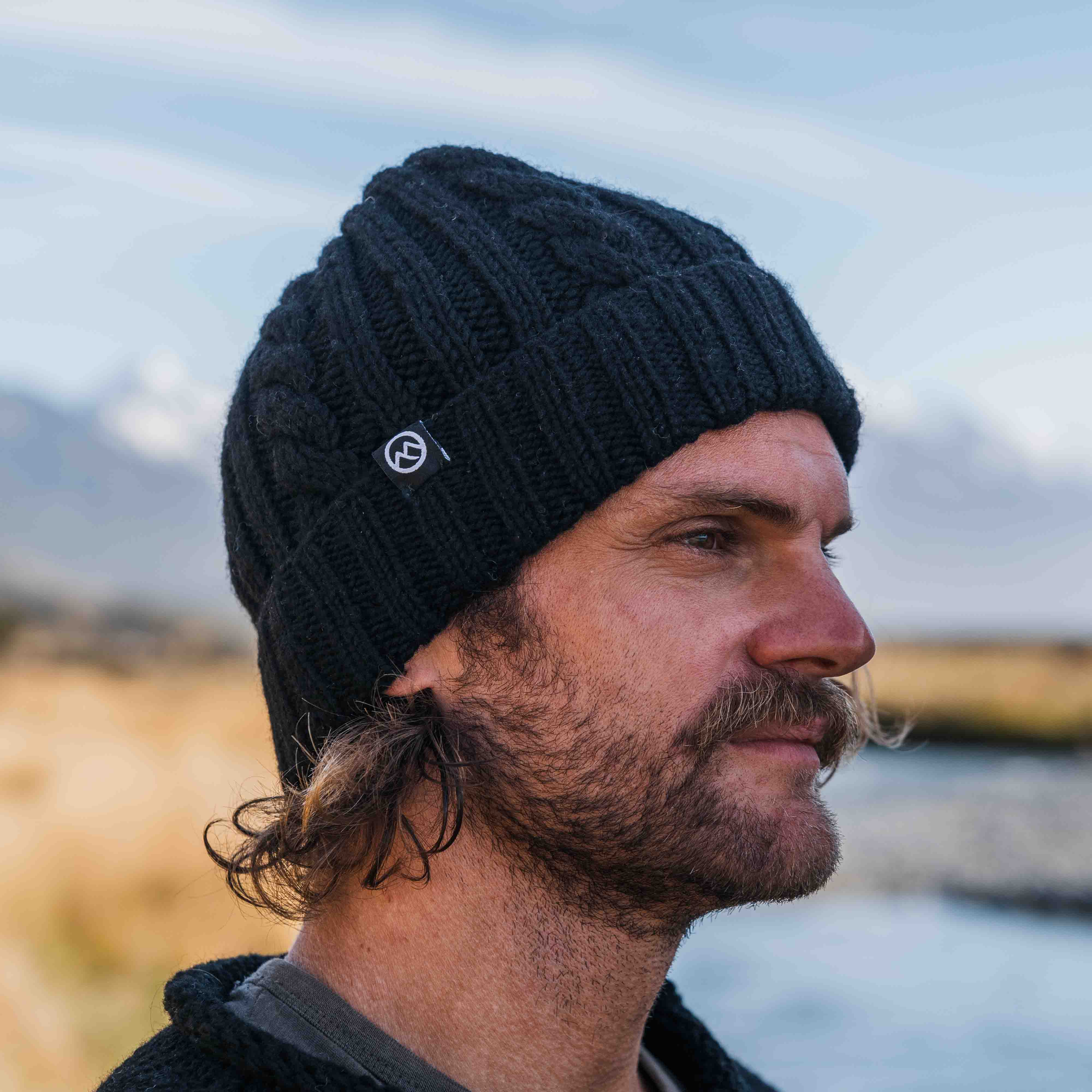 Man wearing a black cable merino wool beanie by Mount Kiwi outdoors near a river with mountains in the background