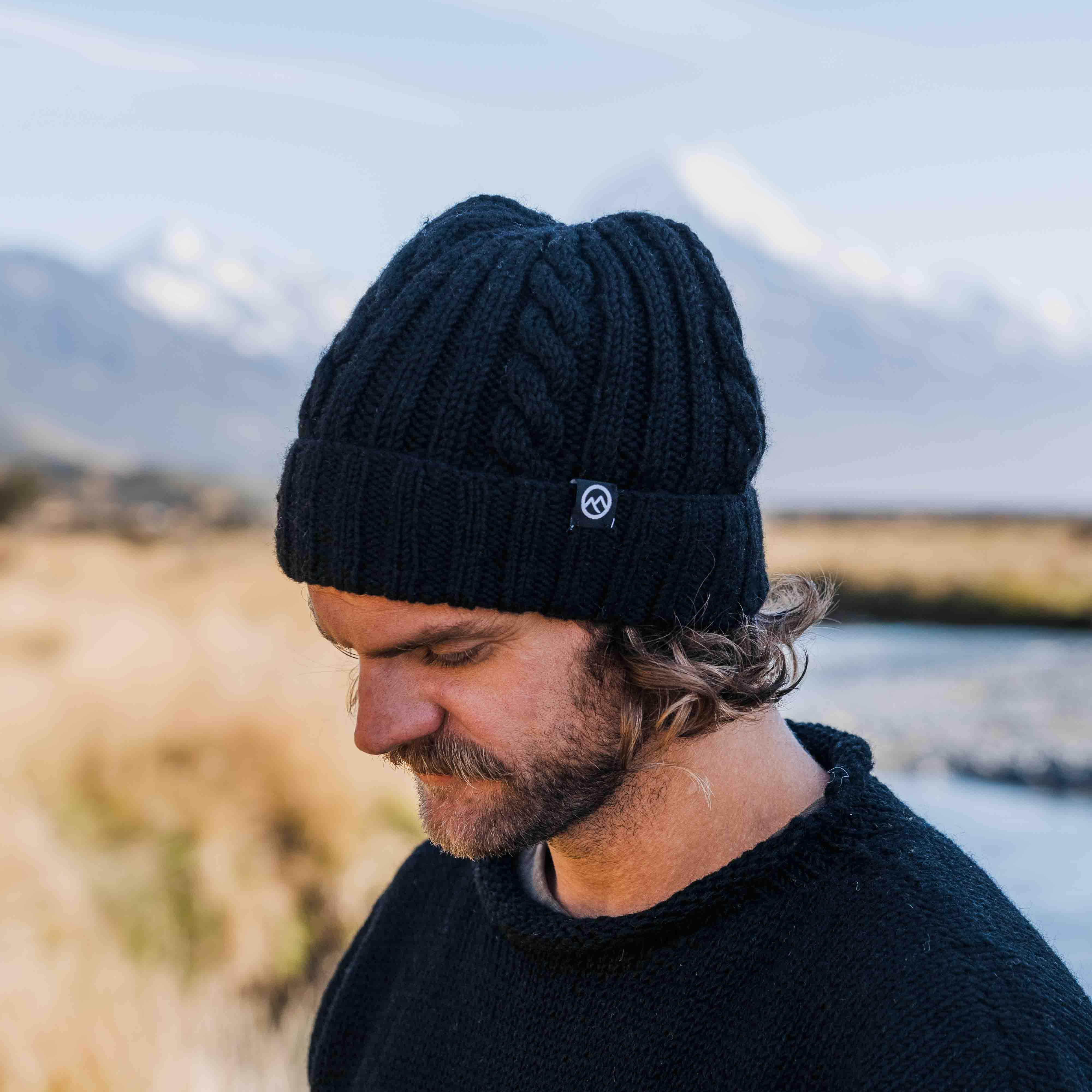 Man wearing a black cable merino wool beanie by Mount Kiwi outdoors near a river with mountains in the background