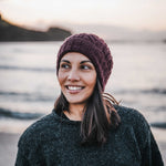 Woman wearing a maroon knit beanie and dark sweater standing outdoors with a blurred natural background