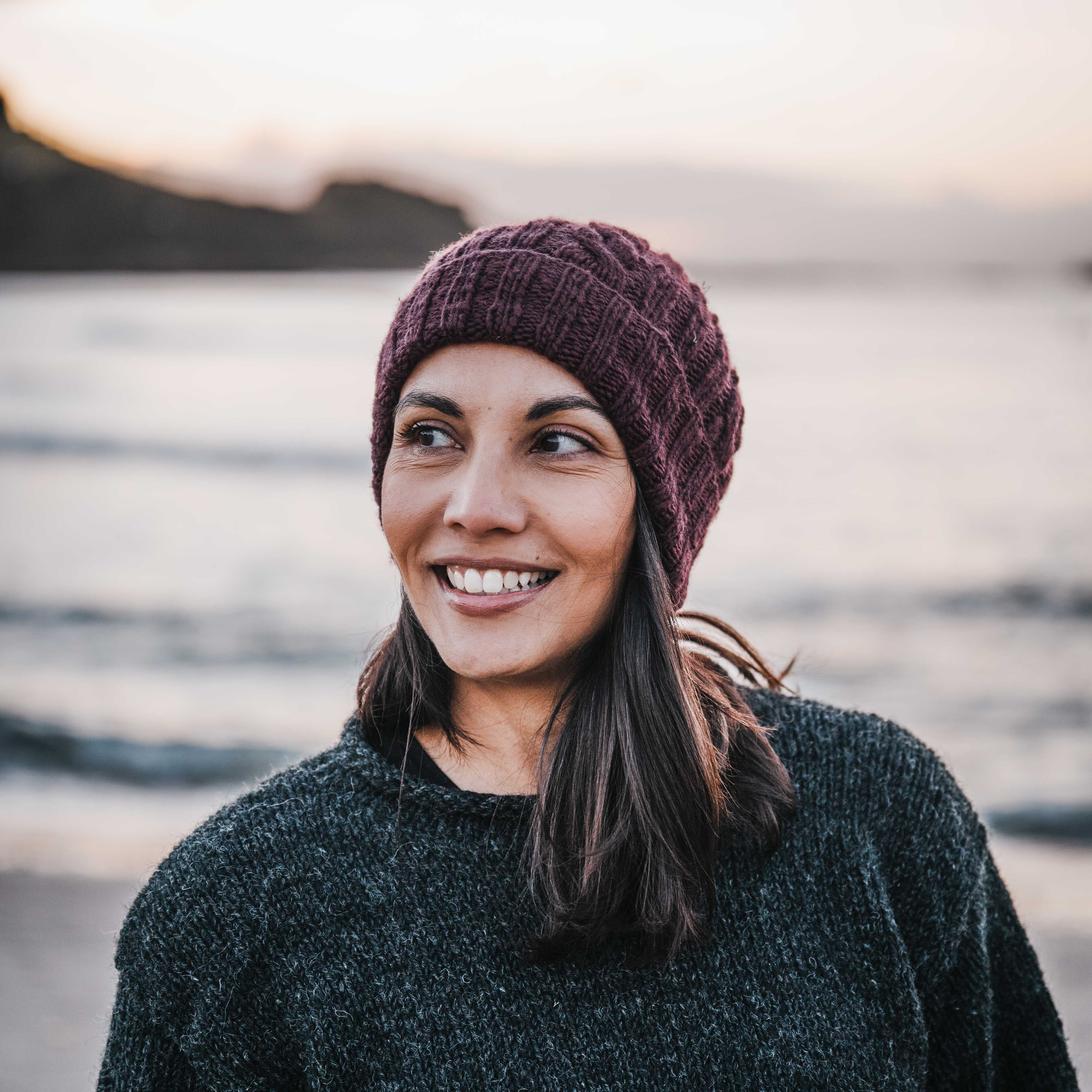 Woman wearing a maroon knit beanie and dark sweater standing outdoors with a blurred natural background